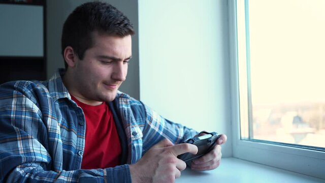 a man playing a small console sitting by the window