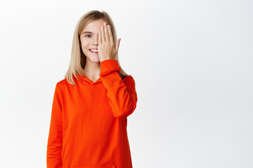 Happy smiling little girl, child covers half of face with palm, looks with one eye at camera, stands in red hoodie against white background