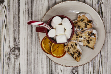 Hot beverage red cup with marshmallows, candy cone stick and Christmas stollen with decorative slices of orange - traditional holidays bread. Festive Christmas background. Christmas tradition food.
