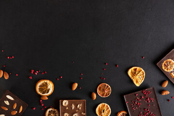 Natural organic milk and bitter chocolate bars with sublimated berries, orange slices, almond and hazelnut on black background. Confectionery ingredients. Copy space.