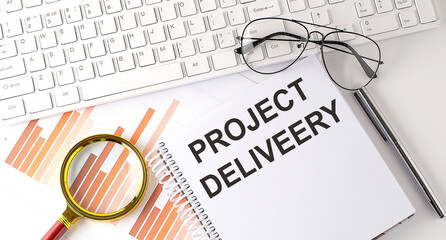 PROJECT DELIVERY text written on a notebook with keyboard, chart,and glasses