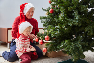 cheerful grandmother and her cute granddaughter girl decorate the Christmas tree. A grandmother and a small child are having fun near a tree indoors.