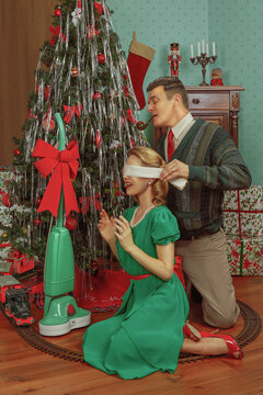 Vintage photography of a married couple from the 1950s in front of a Christmas tree and gifts, the husband removing the blindfold from his wife's eyes to surprise her with a household appliance