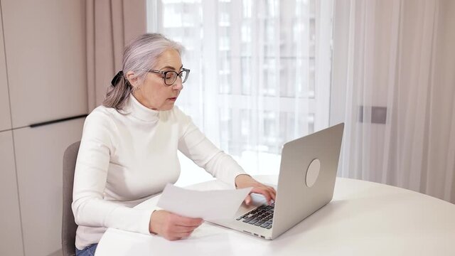 A mature woman works at home on a computer, she looks at documents and works in parallel on a laptop, checking documents