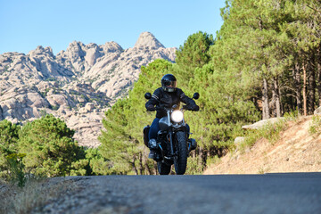 Fototapeta na wymiar Biker in leather jacket and black helmet riding motorbike along hilly road between tall green trees.