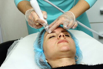 Beauty salon. A cosmetologist in medical gloves and protective mask doing a hydra peeling procedure on the client's cheeks. Side view. Close up. Professional skin care during coronavirus pandemic.