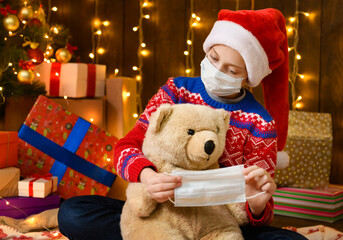 Fototapeta na wymiar Child girl as Santa helper posing in new year decoration. Wearing a protective face mask against viruses, coronaviruses. She puts mask on a toy bear. Holiday lights and lots of gifts