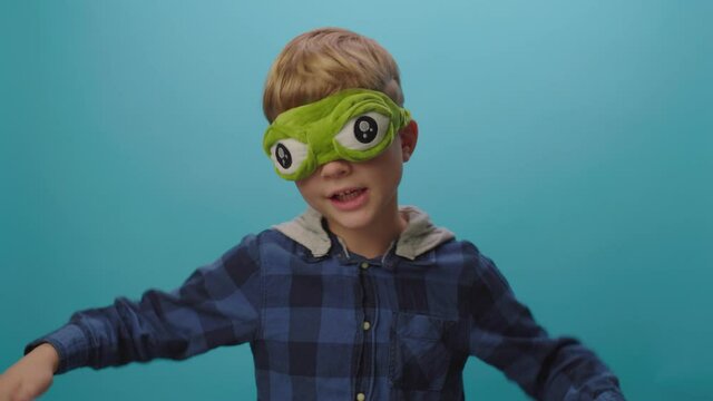 6 years old boy dancing and singing wearing funny frog mask standing on blue background. Kid wearing sleeping mask with big eyes.