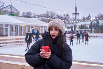 Pretty woman walks around the decorated Christmas city center and drinking coffee on ice rink...