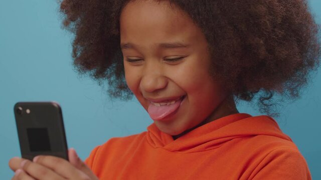 Close up of African American girl making silly faces looking at smartphone camera. Kid is happy to make funny grimaces and show tongue using cell phone. 