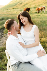 Obraz premium the newlyweds are sitting on a chair and hugging tenderly. girl with a boyfriend on a romantic date at sunset of the day. beautiful couple in the field.