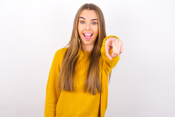 Excited positive Young caucasian girl wearing yellow sweater over white background  points index finger directly at you, sees something very funny. Wow, amazing