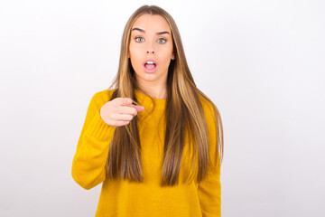 Shocked Young caucasian girl wearing yellow sweater over white background points at you with stunned expression