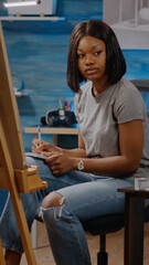 Woman of african american ethnicity writing artistic ideas while sitting in artwork space at home. Black young artist preparing for masterpiece drawing project of authentic white vase