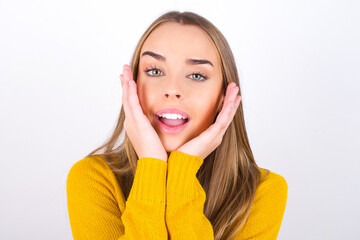 Happy Young caucasian girl wearing yellow sweater over white background touches both cheeks gently, has tender smile, shows white teeth, gazes positively straightly at camera,