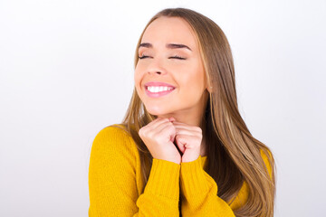 Happy Young caucasian girl wearing yellow sweater over white background anticipates something awesome happen, looks happily aside, keeps hands together near face, has glad expression.