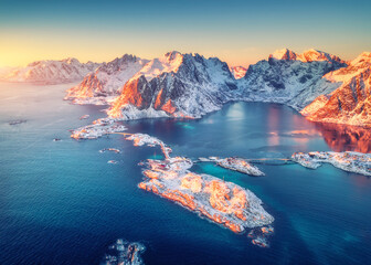 Fototapeta na wymiar Aerial view of snowy islands with rorbu, sea, rocks in water and mountains at sunrise in winter. Beautiful landscape with village, road, bridge. Top view from drone. Hamnoy, Lofoten islands, Norway