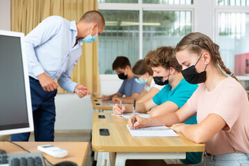 Fototapeta premium Pupils in face masks sitting in class and listening carefully to male teacher.