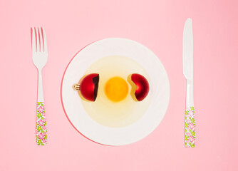 New Year creative layout with plate, egg and red Christmas bauble decoration. Pastel pink background. Holiday party dinner idea.