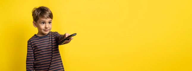 SMall caucasian boy holding tv remote controller while standing in front of yellow background changing channel with copy space - obrazy, fototapety, plakaty
