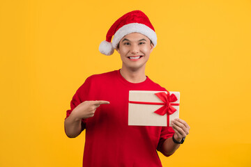 Excited asian handsome man wearing red t-shirt and christmas hat posing isolated on orange background. Mock up copy space. He pointing index finger on gift certificate