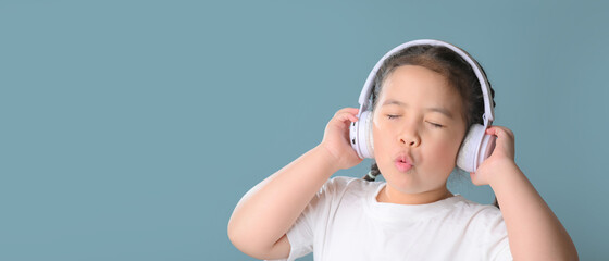 Panoramic shot of happy cute little asian girl  listening music in headphones over blue background. music, technology and people concept.
