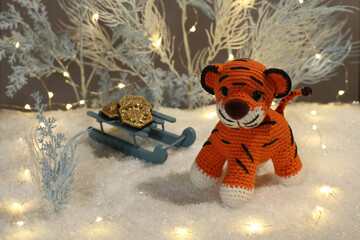 A knitted woolen tiger cub sits next to toy light blue trees with lights and toy sleds with coins on artificial white snow on a gray background. Closeup