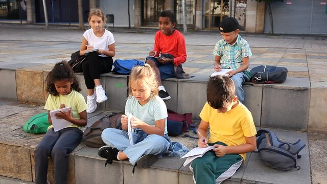 Smiling kids sitting on bench and writing in notepads, studying outdoors 