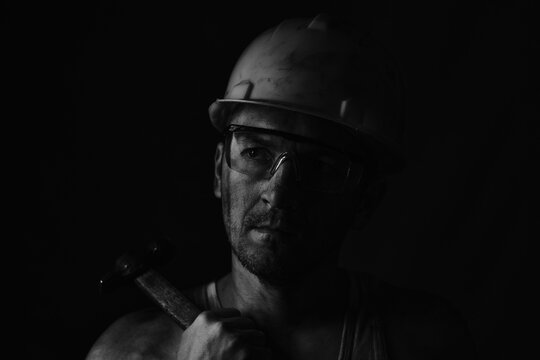 Miner, dirty in coal soot, wearing helmet, protective glasses and with hammer in his hands in black and white photo.