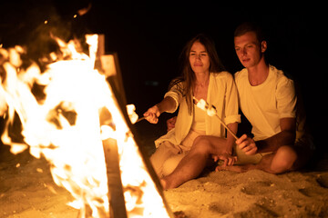 Young couple on the sea beach sitting bonfire and toasting marshmallows on a stick. A romantic date by the fire. Tourism and tourists concept - Powered by Adobe