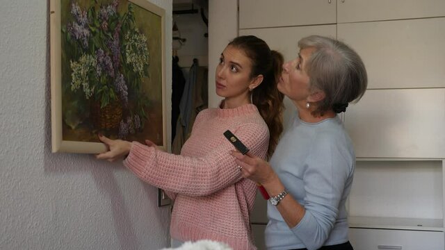 Adult caucasian woman helping her senior grandmother to put painting on wall. Grandmother hammering nail, her daughter standing beside with picture in hands.