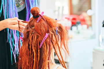 Barber braids dreadlocks. Red-haired girl makes fashionable braids-dreadlocks. Safe removable dreadlocks from kanekalon. Cool hairstyle made of artificial hair. Hairstyle of rainbow Afro-pigtails.