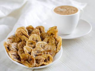 Crispy homemade Banana dried sliced fruits heap on ceramic plate and cup of coffee close up. Healthy snack on white background, side view