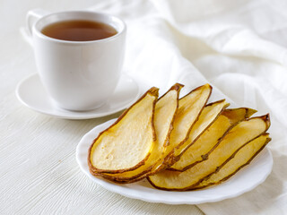 Crispy homemade Pear dried sliced fruits heap on ceramic plate and cup of tea close up. Healthy snack on white background, side view
