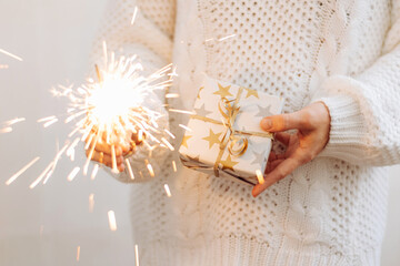 A girl in a white sweater holds a gift and sparkling sparklers. Holiday concept New Year or Christmas. Selective focus