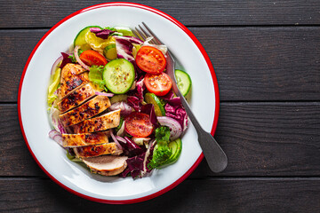 Chicken breast salad with cucumber, tomato and onion in white plate, dark wooden background, top view.