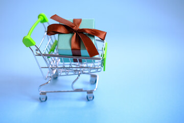 A present box with a ribbon in a shopping trolley cart on blue background . Concept - buying a gift ,online and sale. Christmas, valentines ,women day .Copy space, mockup.