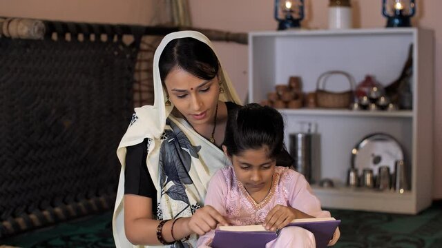 A young lady and her little daughter from a village reading a book together. Pretty female in a printed white Saree and her adorable kid sitting together in the kitchen - rural household  happy par...