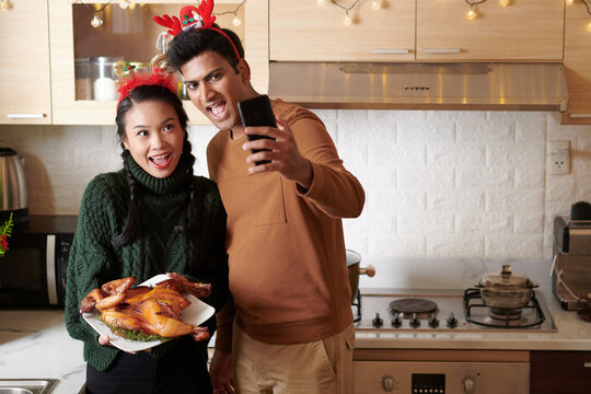 Excited joyful young couple video calling friends or family memebers and showing grilled chicken for Christmas dinner