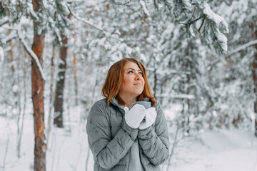 Fototapeta na wymiar A beautiful girl in a gray jacket and knitted white mittens drinks a drink in a snowy forest, looks up and dreams. Comfort and warmth in the winter season