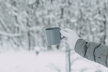 A hand in a white knitted woolen mitt and a gray jacket holds a gray mug against the background of...