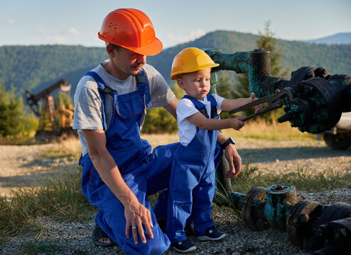Adorable little boy in construction helmet tightening bolt on pipe with industrial wrench while working with father in oil field. Male worker watching how cute child repairing pump jack pipe.