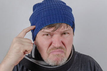A portrait of an angry adult male against a bright wall. Adult man in a blue hat with index finger pointing to his temple.