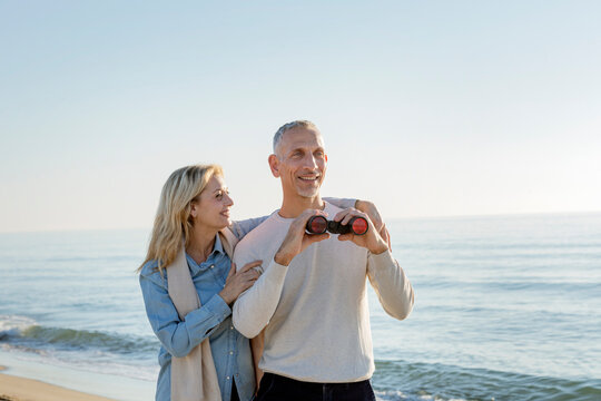 Smiling woman with arm around husband holding binoculars at beach