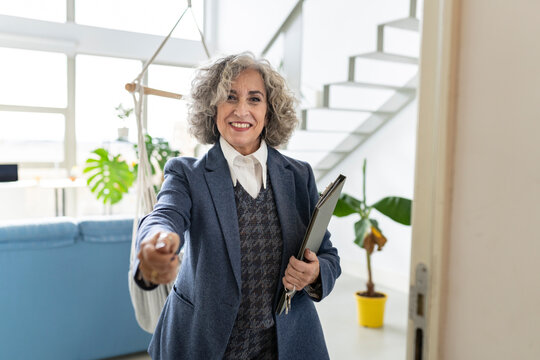 Smiling senior saleswoman extending hand while standing on doorway at apartment