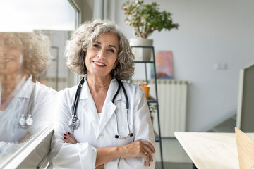 Smiling senior doctor with arms crossed leaning by window in home office