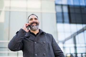 Portrait of happy businessman in black clothing standing in front of company building. Black bearded man is wearing winter clothing and talking on phone.