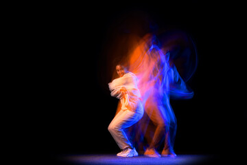 Portrait of young dark skinned flexible girl in white hoodie dancing hip-hop isolated on dark background in purple neon mixed light.