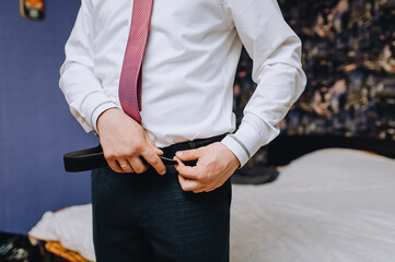 A man, a groom in a white shirt and a red tie puts a leather belt on his trousers, getting ready for a wedding, a wedding in the morning.