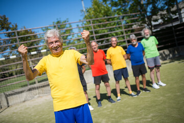 Obraz premium Portrait of happy senior man on football field. Captain with gray hair in sport clothes standing, looking at camera, rising hands, teammates in background. Football, sport, leisure concept